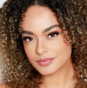 Female model with curly brown hair wearing Reese Robert Beauty's Faux Fur 2D Feather Effect lashes on light gray background